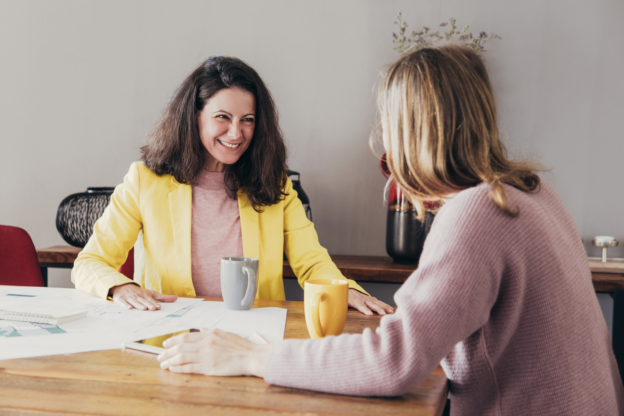 Consultation question image showing two women in warm-toned clothing sitting at a wooden table with coffee mugs and paperwork, engaged in friendly conversation in a cozy, softly lit office.