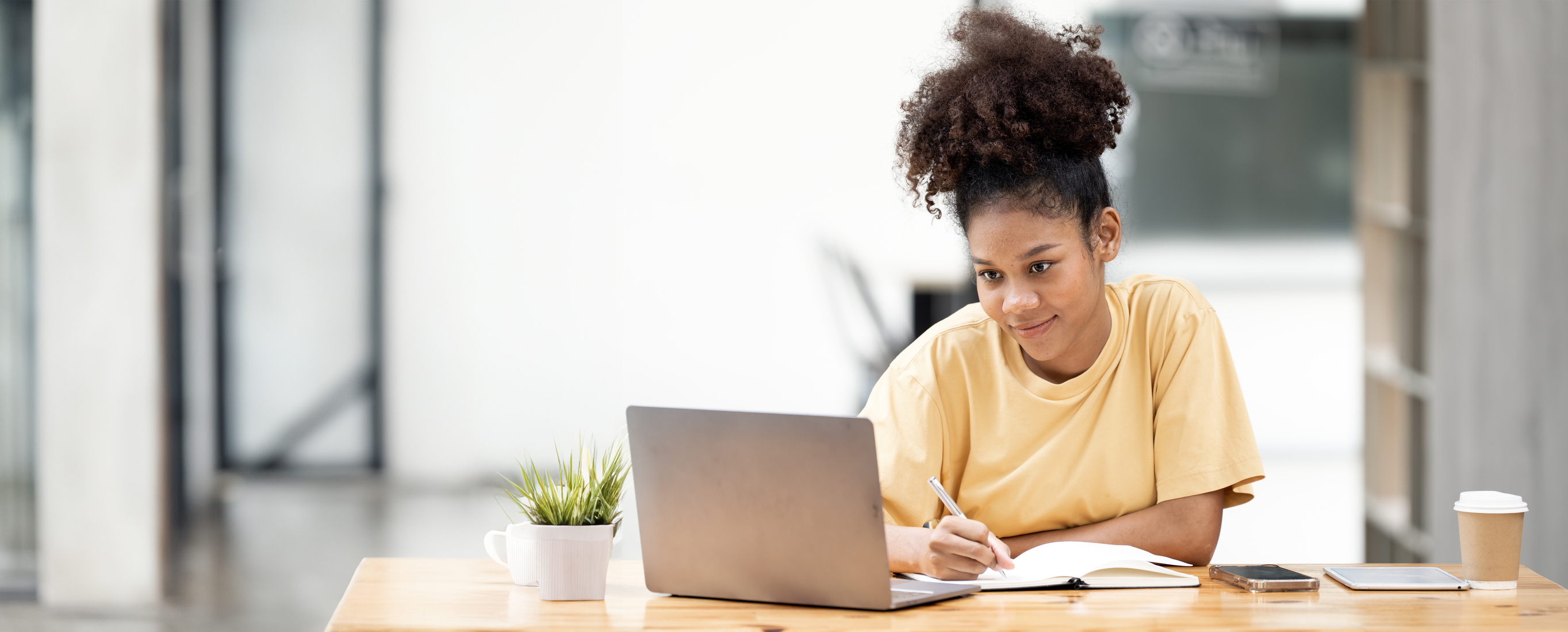 Psychoeducational evaluations image of a student seated at a bright, modern table, writing in a notebook while using a laptop, with school supplies and a small plant nearby.